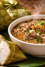 Sajian otentik Coto Makassar dengan pendamping wajib ketupat daun pandan yang wangi dan kenyal di atas meja kayu tradisional.