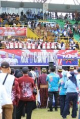 Pemandangan dari belakang kerumunan besar penonton yang berdiri di tribun stadion sepak bola yang penuh di Bone, dengan spanduk "RISE OF CHAMPIONS" di latar belakang.