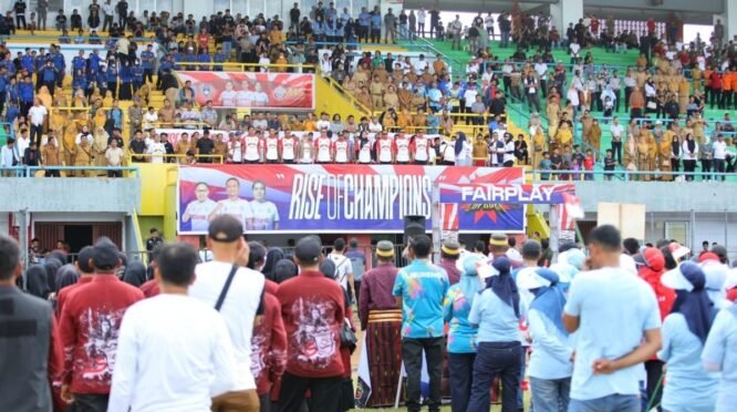 Pemandangan dari belakang kerumunan besar penonton yang berdiri di tribun stadion sepak bola yang penuh di Bone, dengan spanduk "RISE OF CHAMPIONS" di latar belakang.