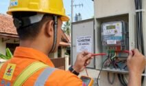 Foto close-up dari arah belakang bahu petugas PLN yang mengenakan seragam oranye dan helm keselamatan kuning. Petugas tersebut tampak sedang memeriksa komponen di dalam kotak meteran listrik digital yang terbuka menggunakan alat tes warna merah. Di latar belakang terlihat suasana perumahan warga dengan atap genting cokelat di bawah langit cerah. Pada baju petugas terdapat logo PLN yang jelas namun tanpa atribut kepolisian.