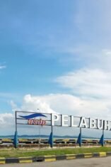 Foto plang nama bertuliskan "PELABUHAN PENYEBERANGAN BAJOE" berwarna putih dengan logo ASDP di sebelah kirinya. Plang ini berdiri di atas beton di tepi laut dengan air laut berwarna kebiruan dan langit biru cerah berawan di latar belakang. Beberapa pohon kelapa dan pohon rindang terlihat di sekitar area plang nama tersebut.