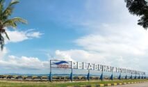 Foto plang nama bertuliskan "PELABUHAN PENYEBERANGAN BAJOE" berwarna putih dengan logo ASDP di sebelah kirinya. Plang ini berdiri di atas beton di tepi laut dengan air laut berwarna kebiruan dan langit biru cerah berawan di latar belakang. Beberapa pohon kelapa dan pohon rindang terlihat di sekitar area plang nama tersebut.