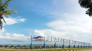 Foto plang nama bertuliskan "PELABUHAN PENYEBERANGAN BAJOE" berwarna putih dengan logo ASDP di sebelah kirinya. Plang ini berdiri di atas beton di tepi laut dengan air laut berwarna kebiruan dan langit biru cerah berawan di latar belakang. Beberapa pohon kelapa dan pohon rindang terlihat di sekitar area plang nama tersebut.