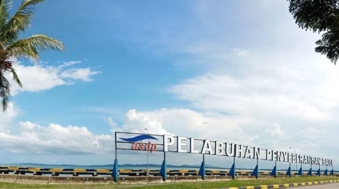 Foto plang nama bertuliskan "PELABUHAN PENYEBERANGAN BAJOE" berwarna putih dengan logo ASDP di sebelah kirinya. Plang ini berdiri di atas beton di tepi laut dengan air laut berwarna kebiruan dan langit biru cerah berawan di latar belakang. Beberapa pohon kelapa dan pohon rindang terlihat di sekitar area plang nama tersebut.
