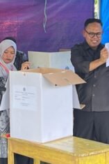 Wakil Bupati Bone, mengenakan kemeja hitam, tampak tersenyum sambil memegang dan menunjukkan selembar surat suara yang menampilkan foto kandidat ke arah depan. Ia berdiri di samping kotak suara berupa kardus putih di atas meja kayu. Di sebelahnya, terdapat panitia perempuan sedang mendampingi. Kegiatan ini berlangsung di atas lapangan berumput di bawah naungan tenda besar berbahan terpal warna biru dan hitam.