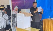 Wakil Bupati Bone, mengenakan kemeja hitam, tampak tersenyum sambil memegang dan menunjukkan selembar surat suara yang menampilkan foto kandidat ke arah depan. Ia berdiri di samping kotak suara berupa kardus putih di atas meja kayu. Di sebelahnya, terdapat panitia perempuan sedang mendampingi. Kegiatan ini berlangsung di atas lapangan berumput di bawah naungan tenda besar berbahan terpal warna biru dan hitam.