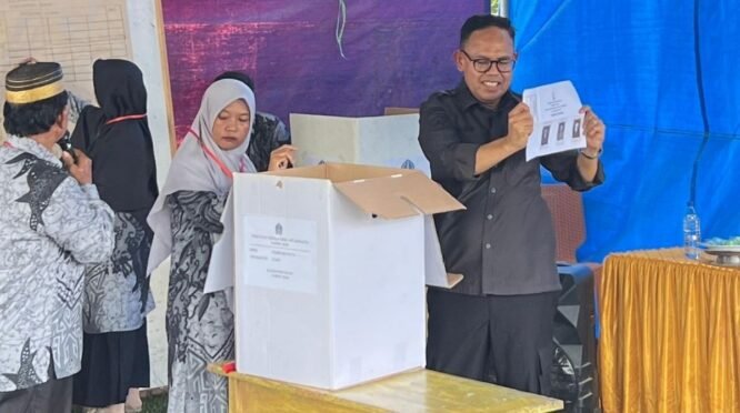 Wakil Bupati Bone, mengenakan kemeja hitam, tampak tersenyum sambil memegang dan menunjukkan selembar surat suara yang menampilkan foto kandidat ke arah depan. Ia berdiri di samping kotak suara berupa kardus putih di atas meja kayu. Di sebelahnya, terdapat panitia perempuan sedang mendampingi. Kegiatan ini berlangsung di atas lapangan berumput di bawah naungan tenda besar berbahan terpal warna biru dan hitam.
