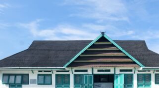 Fasad depan bangunan Museum La Pawawoi Bone dengan arsitektur tradisional atap pelana bertumpuk dan ornamen kepala kerbau di puncaknya, di bawah langit biru berawan.