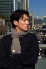 Potret Na Daehoon (Aa Daehoon) mengenakan syal abu-abu sedang berdiri di jembatan penyeberangan jalan dengan latar belakang gedung bertingkat dan Namsan Tower di Seoul, Korea Selatan