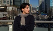 Potret Na Daehoon (Aa Daehoon) mengenakan syal abu-abu sedang berdiri di jembatan penyeberangan jalan dengan latar belakang gedung bertingkat dan Namsan Tower di Seoul, Korea Selatan