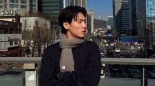 Potret Na Daehoon (Aa Daehoon) mengenakan syal abu-abu sedang berdiri di jembatan penyeberangan jalan dengan latar belakang gedung bertingkat dan Namsan Tower di Seoul, Korea Selatan