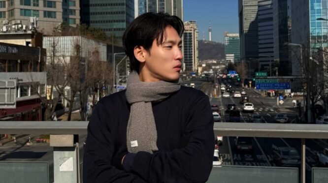 Potret Na Daehoon (Aa Daehoon) mengenakan syal abu-abu sedang berdiri di jembatan penyeberangan jalan dengan latar belakang gedung bertingkat dan Namsan Tower di Seoul, Korea Selatan