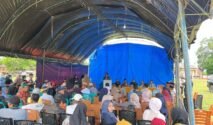 Ratusan warga desa dengan pakaian kasual sedang duduk di deretan kursi plastik di bawah sebuah tenda besar beratap terpal hitam dan biru yang didirikan di atas lapangan berumput. Di bagian depan menghadap ke warga, tampak sejumlah pejabat dan panitia duduk berjajar di belakang meja panjang bertaplak kuning, sementara seorang pria sedang berdiri memberikan arahan melalui podium. Terdapat mobil hitam terparkir di sisi kanan lapangan di bawah langit cerah berawan.