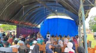 Ratusan warga desa dengan pakaian kasual sedang duduk di deretan kursi plastik di bawah sebuah tenda besar beratap terpal hitam dan biru yang didirikan di atas lapangan berumput. Di bagian depan menghadap ke warga, tampak sejumlah pejabat dan panitia duduk berjajar di belakang meja panjang bertaplak kuning, sementara seorang pria sedang berdiri memberikan arahan melalui podium. Terdapat mobil hitam terparkir di sisi kanan lapangan di bawah langit cerah berawan.