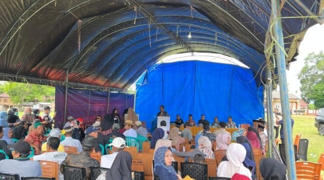 Ratusan warga desa dengan pakaian kasual sedang duduk di deretan kursi plastik di bawah sebuah tenda besar beratap terpal hitam dan biru yang didirikan di atas lapangan berumput. Di bagian depan menghadap ke warga, tampak sejumlah pejabat dan panitia duduk berjajar di belakang meja panjang bertaplak kuning, sementara seorang pria sedang berdiri memberikan arahan melalui podium. Terdapat mobil hitam terparkir di sisi kanan lapangan di bawah langit cerah berawan.