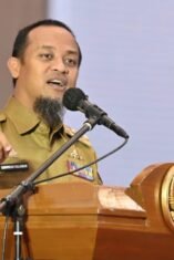 Gubernur Sulawesi Selatan Andi Sudirman Sulaiman berdiri menyampaikan pidato di depan podium resmi dengan latar belakang layar presentasi grafik pertumbuhan makroekonomi daerah. Beliau mengenakan pakaian dinas harian formal, berekspresi optimis saat menjelaskan data rilis BPS terkait PDRB Sulsel triwulan pertama 2026.