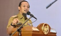 Gubernur Sulawesi Selatan Andi Sudirman Sulaiman berdiri menyampaikan pidato di depan podium resmi dengan latar belakang layar presentasi grafik pertumbuhan makroekonomi daerah. Beliau mengenakan pakaian dinas harian formal, berekspresi optimis saat menjelaskan data rilis BPS terkait PDRB Sulsel triwulan pertama 2026.