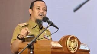 Gubernur Sulawesi Selatan Andi Sudirman Sulaiman berdiri menyampaikan pidato di depan podium resmi dengan latar belakang layar presentasi grafik pertumbuhan makroekonomi daerah. Beliau mengenakan pakaian dinas harian formal, berekspresi optimis saat menjelaskan data rilis BPS terkait PDRB Sulsel triwulan pertama 2026.