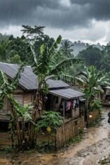 Pemandangan udara rendah dari sebuah perkampungan sederhana di pedesaan saat hujan deras. Di tengah cuaca mendung gelap dan rintik hujan yang terlihat jelas, beberapa rumah kayu sederhana dengan atap seng berjejer di antara pepohonan rimbun, termasuk pohon-pohon pisang yang basah. Seorang warga mengenakan jas hujan tampak sedang berjalan melintasi jalan tanah yang becek dan penuh genangan air.