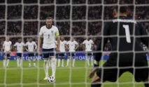 Harry Kane mengenakan seragam Timnas Inggris bersiap mengambil tendangan penalti dengan sudut pandang dari belakang gawang lawan di stadion sepak bola.