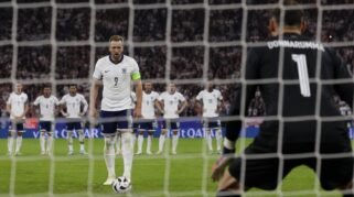 Harry Kane mengenakan seragam Timnas Inggris bersiap mengambil tendangan penalti dengan sudut pandang dari belakang gawang lawan di stadion sepak bola.